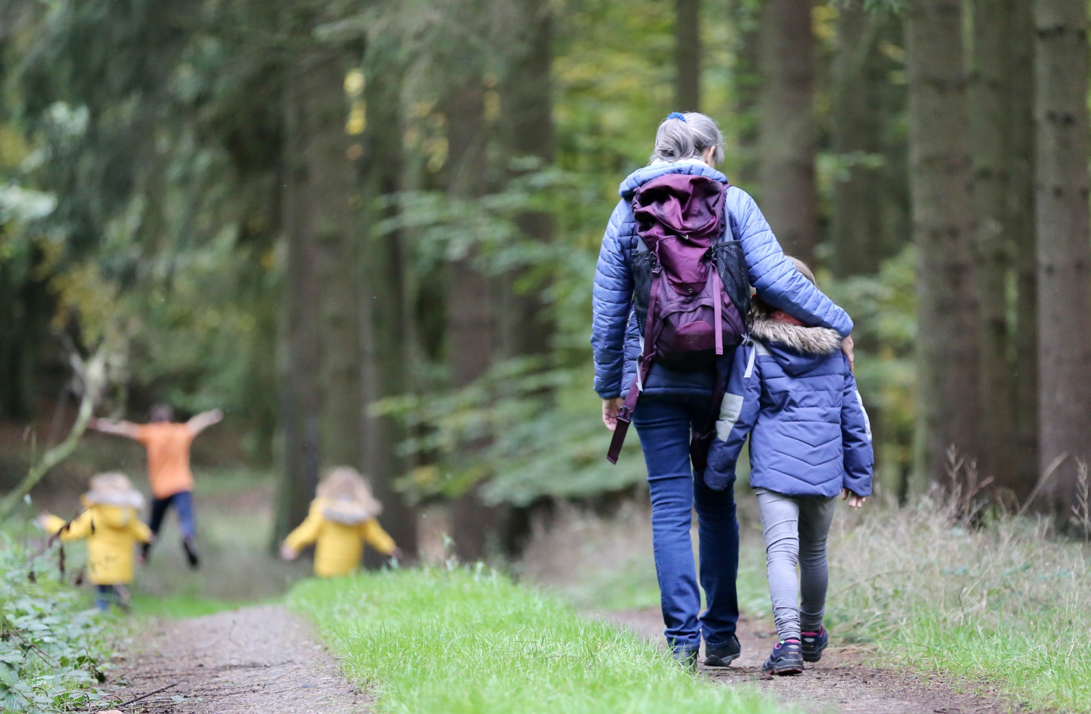 mom and daughter walking in the woods with arms around each other while kids play in the distance